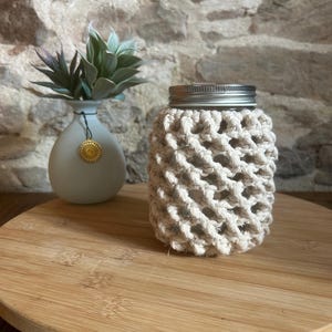 Peut inclure: Un bocal en verre enveloppé d'une corde en macramé couleur crème avec un motif de nœuds en losange et un couvercle en métal argenté. À côté se trouve un petit vase en céramique gris clair contenant une plante succulente verte avec une breloque dorée.