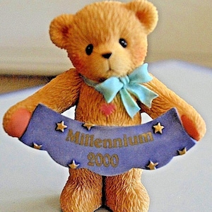 Puede incluir: Un pequeño oso de peluche marrón sosteniendo una pancarta azul con estrellas doradas y el texto "Millennium 2000". El oso lleva un lazo azul claro.