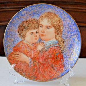 Puede incluir: Un plato de porcelana decorativo con una pintura de una mujer sosteniendo a un niño. La mujer y el niño visten ropa colorida y estampada. El fondo es un diseño floral azul y dorado.