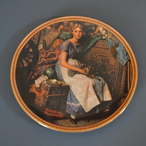 May include: A round ceramic plate with a gold rim features a painting of a young woman sitting on a wooden chest. The woman is wearing a blue dress and a white apron. The painting is surrounded by a decorative border of gold and red.