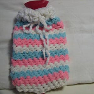 May include: A hot water bottle encased in a hand-crocheted cover featuring horizontal stripes of white, light blue, and pink yarn. The cover includes a white ruffled collar at the top with a matching white drawstring tie and a red stopper visible.