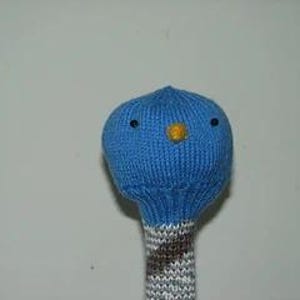 May include: A blue knitted golf club head cover featuring a rounded top with two small dark eyes and a yellow beak. The neck of the cover is made of a ribbed knit fabric with a grey and brown diagonal striped pattern extending downwards.