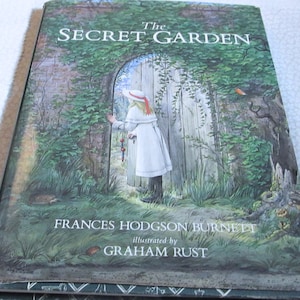 May include: A hardcover book titled "The Secret Garden" with an illustration of a girl in a white dress standing before a wooden door in a garden. The author's name, Frances Hodgson Burnett, and the illustrator's name, Graham Rust, are also visible.
