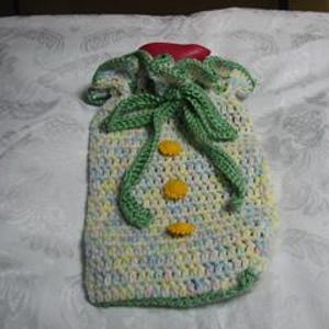 Puede incluir: Una funda para bolsa de agua caliente hecha de hilo pastel jaspeado en tonos azul, amarillo y blanco. La funda presenta un borde superior con volantes, un cordón de ganchillo verde y tres pequeños botones decorativos redondos amarillos.