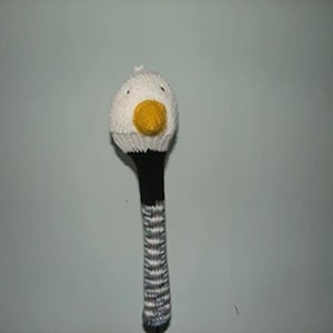 May include: A hand-knitted golf club head cover featuring a white bird head with a yellow beak and small dark eyes. The neck of the cover is solid black, transitioning into a multi-colored striped pattern of white, grey, and blue at the base.