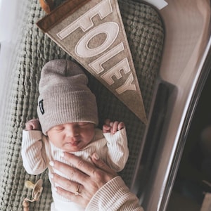 Peut inclure: Un nouveau-n&eacute; portant un bonnet beige et un ensemble c&ocirc;tel&eacute; assorti, couch&eacute; sur une couverture verte textur&eacute;e. Une banni&egrave;re en feutre avec le nom "FOLEY" est visible. Une sucette avec une cha&icirc;ne de perles en bois est &eacute;galement pr&eacute;sente.