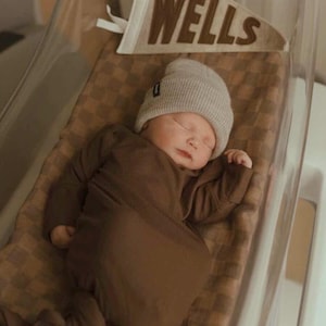 Peut inclure: Un nouveau-n&eacute; endormi, v&ecirc;tu d'une tenue marron et d'un bonnet gris, dans un berceau transparent. Une banni&egrave;re avec le nom "WELLS" est visible au-dessus du b&eacute;b&eacute;. Le berceau est tapiss&eacute; d'une couverture marron &agrave; carreaux.