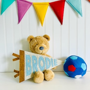 Peut inclure: Un ours en peluche marron tient une banni&egrave;re en feutre avec le nom "BRODIE" en lettres bleu clair. Un ballon de football bleu et rouge est &agrave; droite. Des banni&egrave;res triangulaires en feutre color&eacute;es sont suspendues en arri&egrave;re-plan.