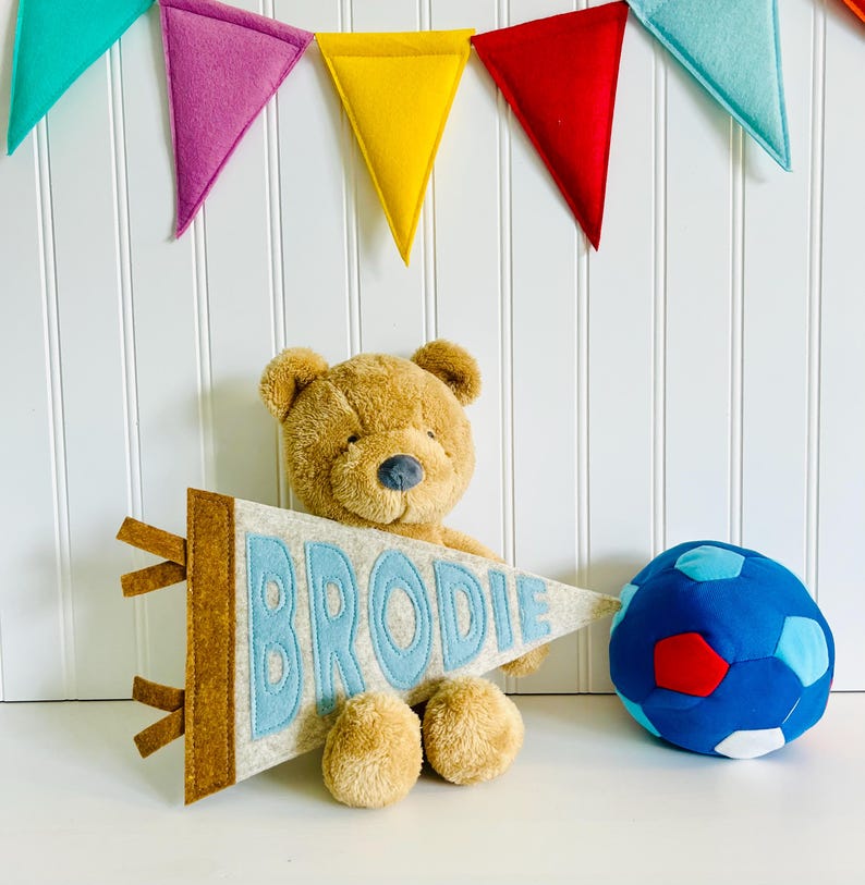 Peut inclure: Un ours en peluche marron tient une banni&egrave;re en feutre avec le nom "BRODIE" en lettres bleu clair. Un ballon de football bleu et rouge est &agrave; droite. Des banni&egrave;res triangulaires en feutre color&eacute;es sont suspendues en arri&egrave;re-plan.