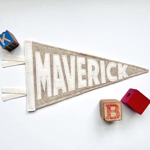 Peut inclure: Une banni&egrave;re en feutre beige avec le nom "MAVERICK" en lettres blanches. La banni&egrave;re a une bordure et des glands blancs. Des blocs de lettres en bois avec les lettres "K" et "B" sont &eacute;galement dans l'image.