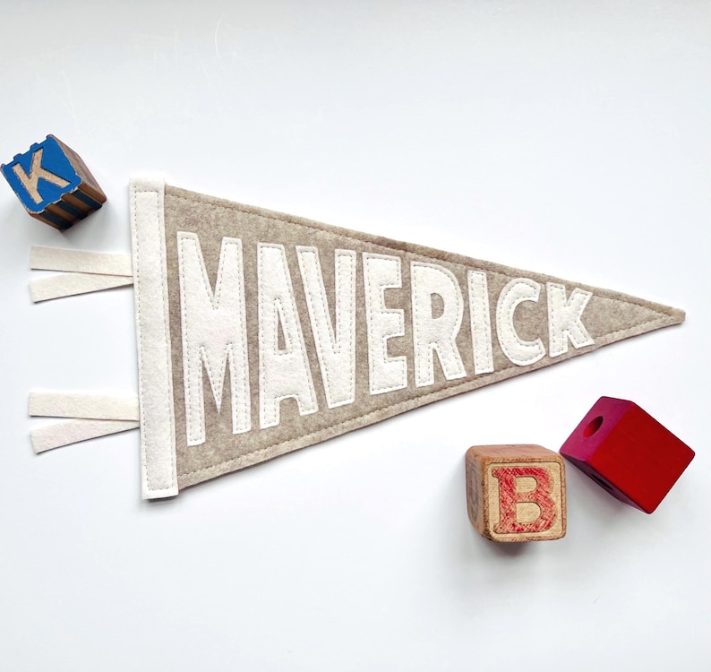 Peut inclure: Une banni&egrave;re en feutre beige avec le nom "MAVERICK" en lettres blanches. La banni&egrave;re a une bordure et des glands blancs. Des blocs de lettres en bois avec les lettres "K" et "B" sont &eacute;galement dans l'image.