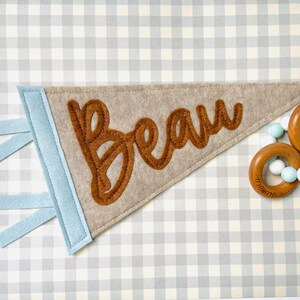 May include: A beige felt pennant with the name "Beau" in brown script. The pennant has a light blue border and matching hanging ribbons. A wooden teething ring with blue and white beads is in the background. The background is a gray and white checkered pattern.