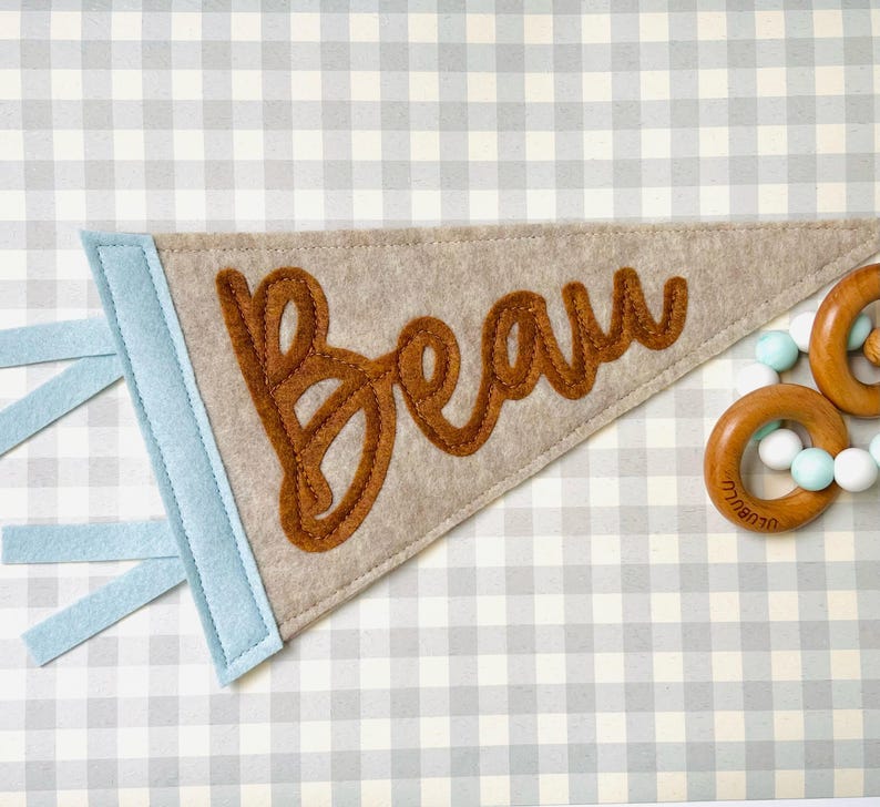 May include: A beige felt pennant with the name "Beau" in brown script. The pennant has a light blue border and matching hanging ribbons. A wooden teething ring with blue and white beads is in the background. The background is a gray and white checkered pattern.