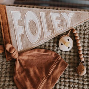 Peut inclure: Un fanion en feutre gris clair avec le nom "FOLEY" en lettres blanches, un bonnet nou&eacute; marron et une sucette avec une cha&icirc;ne de perles en bois. Articles pour b&eacute;b&eacute;s.