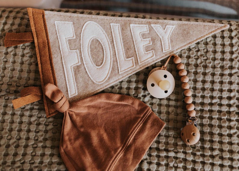 Peut inclure: Un fanion en feutre gris clair avec le nom "FOLEY" en lettres blanches, un bonnet nou&eacute; marron et une sucette avec une cha&icirc;ne de perles en bois. Articles pour b&eacute;b&eacute;s.