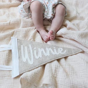 May include: A beige felt pennant with the name "Winnie" in white script. The pennant has a white border and white fabric loops for hanging. The pennant is lying on a beige blanket, with a baby's legs and feet visible above.