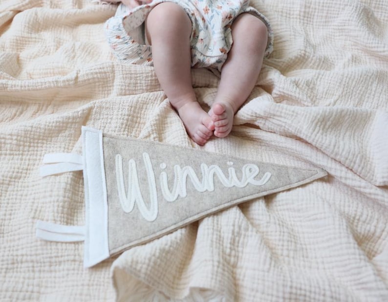 May include: A beige felt pennant with the name "Winnie" in white script. The pennant has a white border and white fabric loops for hanging. The pennant is lying on a beige blanket, with a baby's legs and feet visible above.