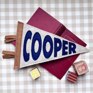 Peut inclure: Un fanion personnalis&eacute; avec le nom "COOPER" en lettres bleues sur un fond en feutre gris. Le fanion a une bordure marron et une frange en cuir. Un livre bordeaux et des blocs de bois avec des lettres sont &eacute;galement dans l'image.
