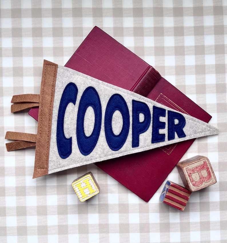 Peut inclure: Un fanion personnalis&eacute; avec le nom "COOPER" en lettres bleues sur un fond en feutre gris. Le fanion a une bordure marron et une frange en cuir. Un livre bordeaux et des blocs de bois avec des lettres sont &eacute;galement dans l'image.