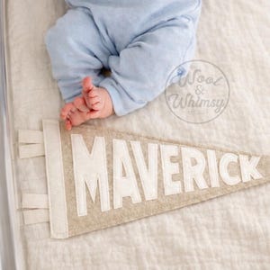 Peut inclure: Une banni&egrave;re en feutre beige avec le nom "MAVERICK" en lettres blanches. La banni&egrave;re a une bordure blanche et est pos&eacute;e sur une surface blanche. Un b&eacute;b&eacute; en barboteuse bleue est allong&eacute; &agrave; c&ocirc;t&eacute; de la banni&egrave;re.