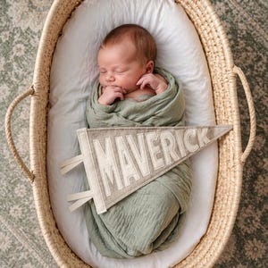 Peut inclure: Un nouveau-n&eacute; endormi, envelopp&eacute; dans une couverture vert sauge, repose dans un panier en osier. Une banni&egrave;re avec le nom "MAVERICK" est plac&eacute;e pr&egrave;s du b&eacute;b&eacute;. Le panier est garni d'un drap blanc.