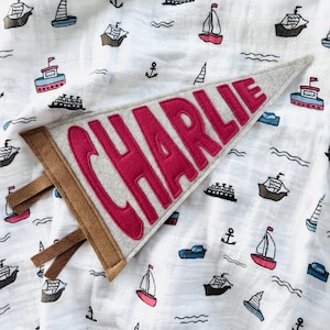 Peut inclure: Une banni&egrave;re en feutre gris avec le nom "CHARLIE" en lettres rouges. La banni&egrave;re a une bordure marron et des lani&egrave;res de tissu. Elle est pos&eacute;e sur un tissu blanc &agrave; motifs nautiques de bateaux, d'ancres et de vagues.