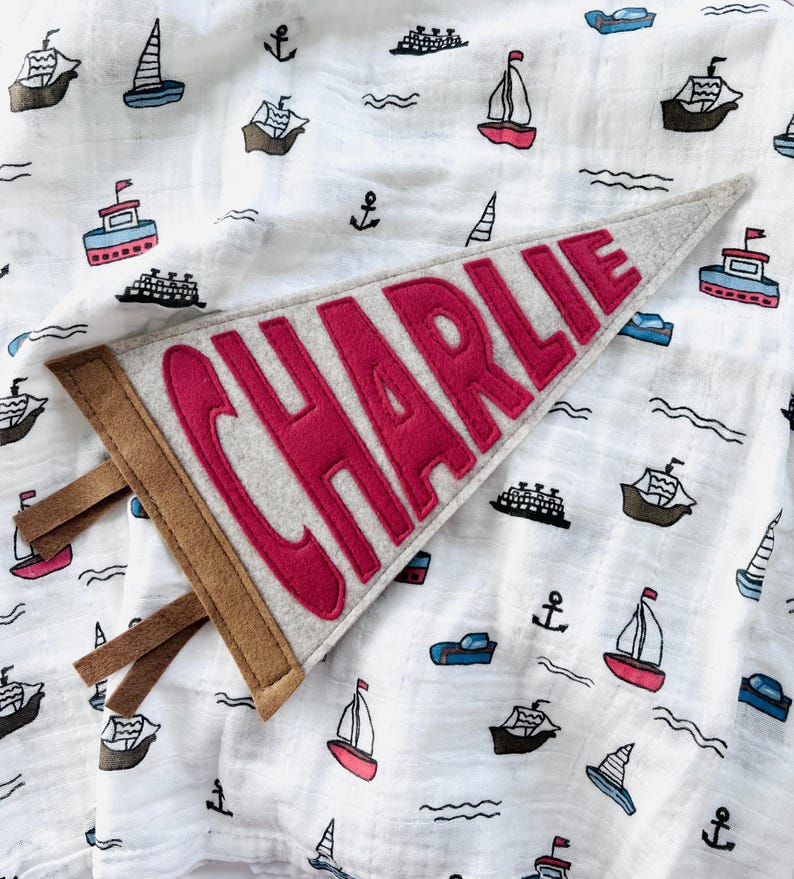 Peut inclure: Une banni&egrave;re en feutre gris avec le nom "CHARLIE" en lettres rouges. La banni&egrave;re a une bordure marron et des lani&egrave;res de tissu. Elle est pos&eacute;e sur un tissu blanc &agrave; motifs nautiques de bateaux, d'ancres et de vagues.