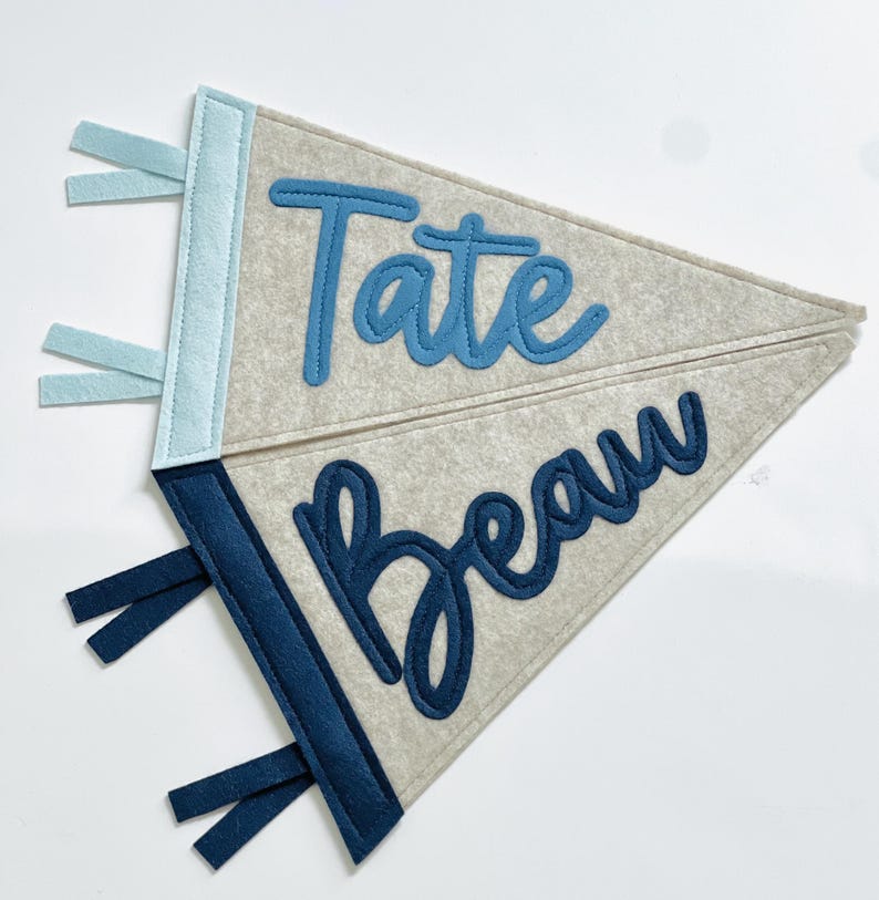 May include: Two gray felt pennants with blue accents. One pennant has the name "Tate" in light blue script, and the other has "Beau" in a darker blue script. Each pennant has a blue border and matching blue ribbons.