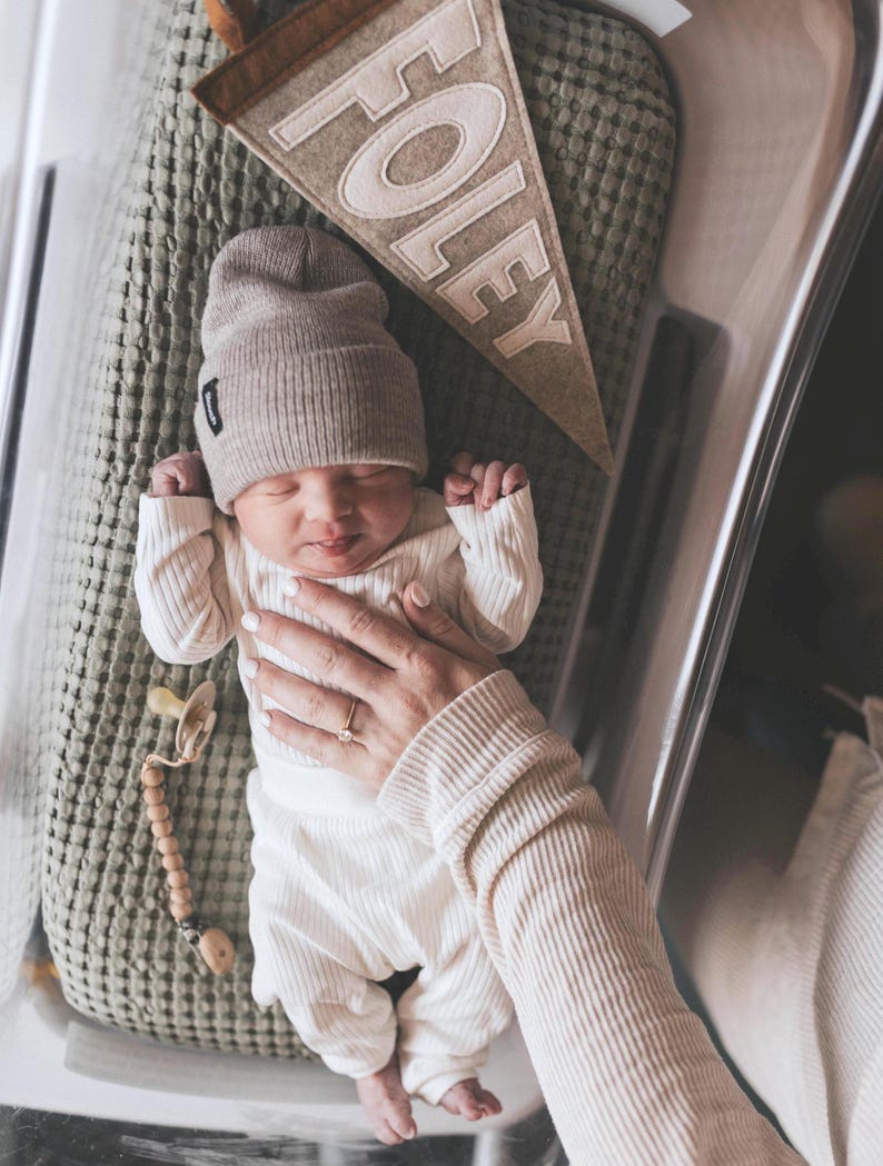 Peut inclure: Un nouveau-n&eacute; portant un bonnet beige et un ensemble c&ocirc;tel&eacute; assorti, couch&eacute; sur une couverture verte textur&eacute;e. Une banni&egrave;re en feutre avec le nom "FOLEY" est visible. Une sucette avec une cha&icirc;ne de perles en bois est &eacute;galement pr&eacute;sente.