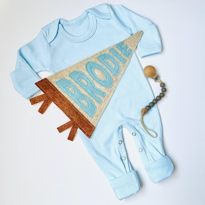 Peut inclure: Un pyjama b&eacute;b&eacute; bleu clair &agrave; manches longues et fermetures &agrave; boutons-pression. Un fanion en feutre avec le nom "BRODIE" en lettres bleu clair est plac&eacute; sur le pyjama. Une attache-t&eacute;tine en bois et perles est &eacute;galement incluse.