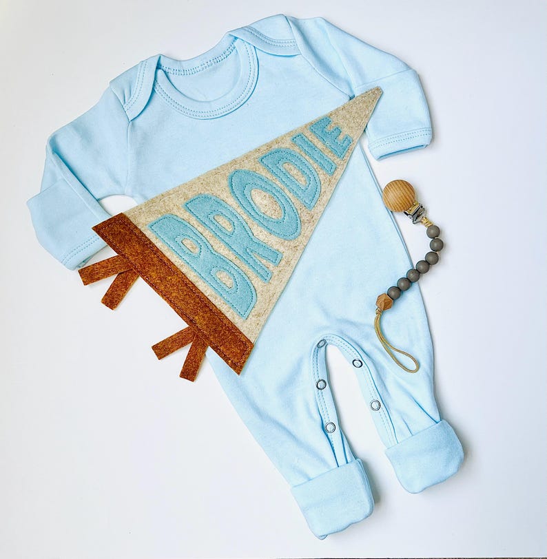 Peut inclure: Un pyjama b&eacute;b&eacute; bleu clair &agrave; manches longues et fermetures &agrave; boutons-pression. Un fanion en feutre avec le nom "BRODIE" en lettres bleu clair est plac&eacute; sur le pyjama. Une attache-t&eacute;tine en bois et perles est &eacute;galement incluse.