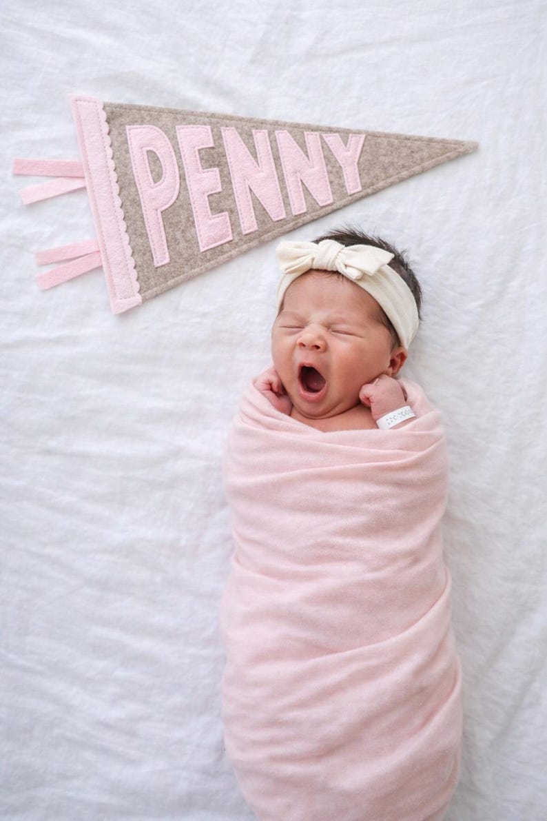 Peut inclure: Un nouveau-n&eacute; envelopp&eacute; dans une couverture rose tendre, b&acirc;illant. Un bandeau avec un n&oelig;ud de couleur cr&egrave;me orne la t&ecirc;te du b&eacute;b&eacute;. Une banni&egrave;re en feutre avec le nom "PENNY" en lettres roses est au-dessus du b&eacute;b&eacute;.