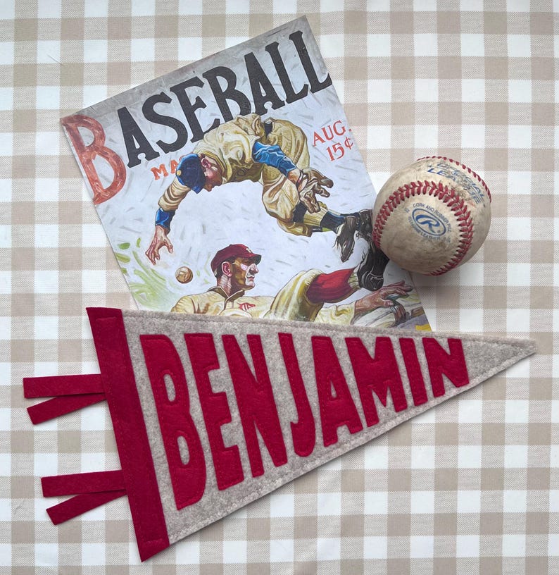 Peut inclure: Une composition sur le th&egrave;me du baseball avec une couverture de magazine vintage, une balle de baseball et une banni&egrave;re en feutre. La banni&egrave;re est grise avec le nom "BENJAMIN" en lettres rouges. La couverture du magazine repr&eacute;sente des joueurs de baseball en action.