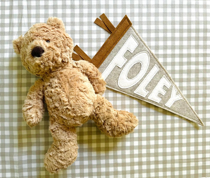 Peut inclure: Un ours en peluche brun clair avec un nez brun fonc&eacute; et une banni&egrave;re grise avec le nom "FOLEY" en lettres blanches. La banni&egrave;re a une bordure marron et est de forme triangulaire. Sur un fond &agrave; carreaux.