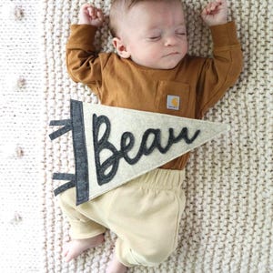 May include: A sleeping baby wearing a brown long-sleeve shirt and tan pants. A felt banner with the name "Beau" in black script is placed near the baby. The baby is lying on a cream-colored knitted blanket.