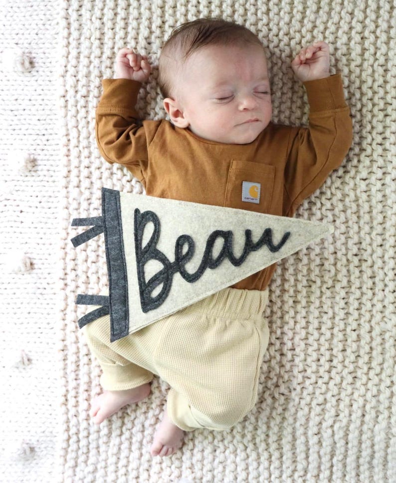 May include: A sleeping baby wearing a brown long-sleeve shirt and tan pants. A felt banner with the name "Beau" in black script is placed near the baby. The baby is lying on a cream-colored knitted blanket.