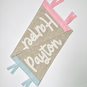 May include: Two felt pennant flags, one with pink trim and the name "Harper" in white script, the other with blue trim and the name "Payton". The pennants are beige and have matching pink and blue ribbon accents.