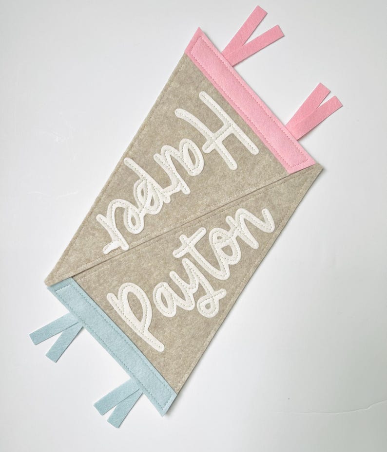 May include: Two felt pennant flags, one with pink trim and the name "Harper" in white script, the other with blue trim and the name "Payton". The pennants are beige and have matching pink and blue ribbon accents.