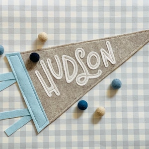 May include: A gray felt pennant with the name "HUDSON" in white script. The pennant has a light blue accent and is decorated with small, colorful felt balls. The background is a light blue and white checkered pattern.