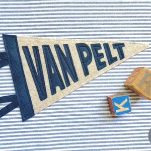 Peut inclure: Une banni&egrave;re en feutre beige avec le nom "VAN PELT" en lettres bleu marine. La banni&egrave;re a une bordure et des attaches bleu marine. Deux blocs de lettres en bois sont &agrave; c&ocirc;t&eacute; de la banni&egrave;re, dont un avec la lettre "K". Le fond est un tissu &agrave; rayures bleues et blanches.