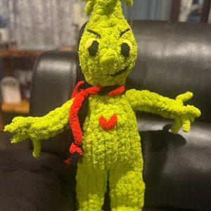 May include: A handmade, green crocheted Grinch doll with black eyes, a red heart, and a red and black scarf. The Grinch has a mischievous grin and is posed with arms outstretched. The doll is approximately 12 inches tall.