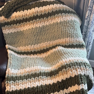 May include: A striped knitted throw blanket in shades of green, white, and beige. The blanket is made of chunky yarn and has a soft, fluffy texture.