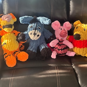 May include: Four handmade plush toys of Winnie the Pooh characters: Tigger, Eeyore, Piglet, and Winnie the Pooh. Tigger is orange and yellow, Eeyore is blue, Piglet is pink, and Winnie the Pooh is yellow with a red shirt. They are sitting on a black surface.