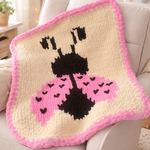 May include: A cream-colored, square crochet blanket with a pink border. The blanket features a large ladybug design in black and pink. The blanket is draped over a white armchair.