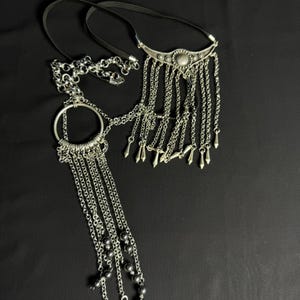 May include: Two silver chain necklaces with decorative pendants. One necklace has a large circular pendant with smaller chains and beads hanging from it. The other necklace has a pointed pendant with smaller chains hanging from it.