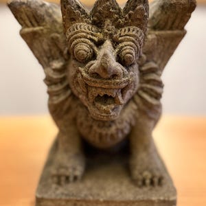 Może przedstawiać: Szczegółowa kamienna rzeźba mitycznego stworzenia ze skrzydłami, koroną i otwartymi ustami. Posąg ma stonowany szary kolor i stoi na prostokątnej podstawie. Stworzenie ma groźny wyraz twarzy.