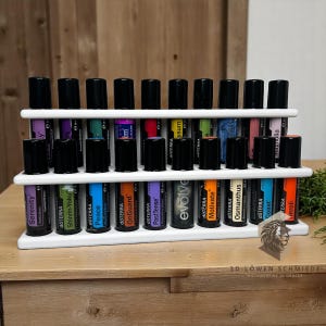 Könnte beinhalten: Ein weißes, mehrstöckiges Display für ätherische Öle mit zahlreichen Flaschen ätherischer Öle mit schwarzen Kappen. Die Flaschen sind mit verschiedenen Namen und Farben beschriftet. Das Regal besteht aus weißem Material.