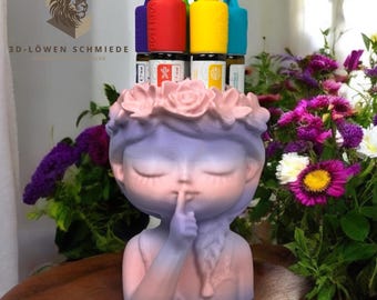 May include: A decorative planter in the shape of a girl's face, with a finger to her lips, painted in shades of pink and purple. The planter is adorned with a floral crown and holds several essential oil bottles with colorful caps. The background features vibrant flowers.