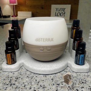 Op de afbeelding: Een witte en bruine etherische olie diffuser met de merknaam "dōTERRA" erop. De diffuser staat op een grijze, gespikkelde tray met verschillende kleine flesjes etherische olie. De tekst "Yu'te Loo!" is zichtbaar op de achtergrond.