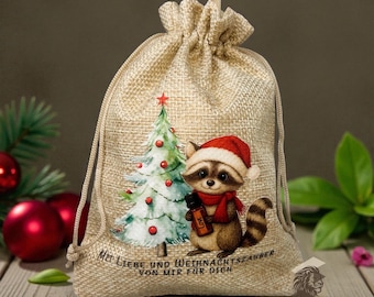 May include: A beige burlap drawstring gift bag featuring a Christmas tree and a raccoon wearing a Santa hat and scarf. The raccoon holds a bottle with the word "Love". The bag has text in German.